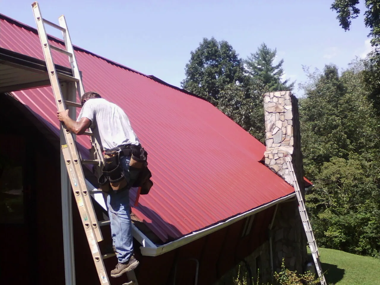 Roofer on a ladder beside a red metal roof and stone chimney — Soffit & Fascia Repair by Roofing Brothers in Whitharral